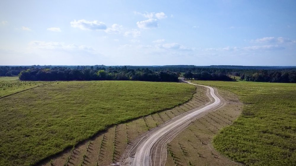 A winding dirt road cuts through a wide, open field under a clear blue sky, surrounded by trees in the distance. The landscape appears ready for construction or land management activities.