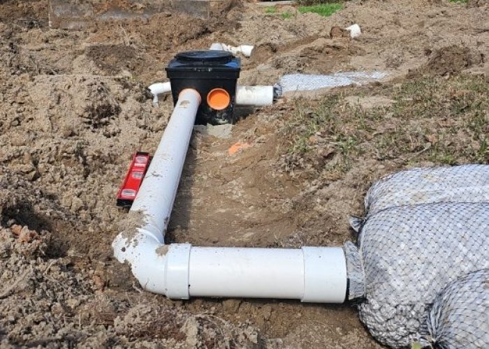 A trench is dug in a yard, showing white PVC pipes leading to a black drainage box, with an excavator in the background. The area is surrounded by bare soil and some grass.