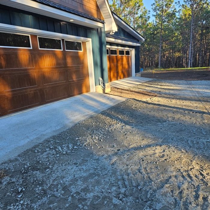Two garage doors with wooden panels are shown next to a gravel driveway, surrounded by a forested area. The sunlight casts shadows on the ground, highlighting the textures of the surfaces.