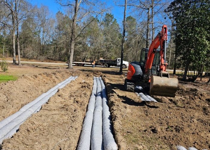 Construction site with an excavator and several gray pipes laid out along a trench. Trees are visible in the background under a clear blue sky.