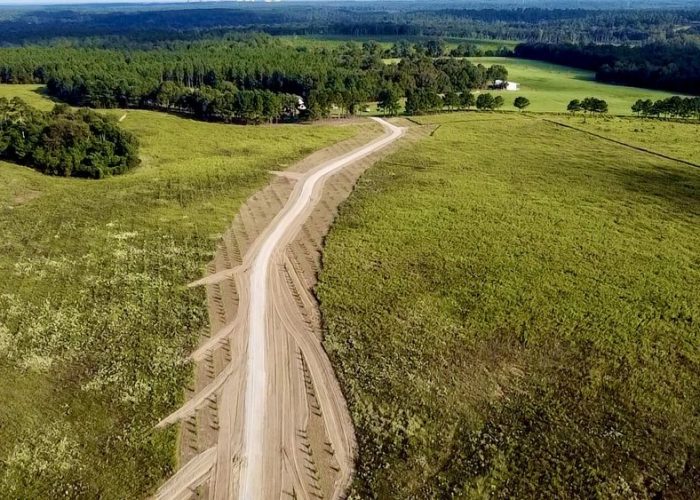 Aerial view of a winding dirt road cutting through a vast, open field surrounded by trees and distant hills. The landscape is predominantly green with patches of grass and earth.