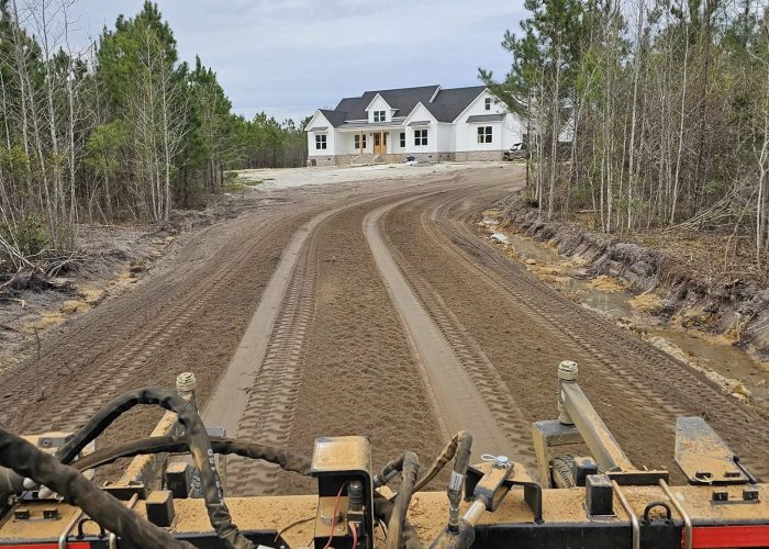 View from a construction vehicle showing a newly graded dirt road leading to a house in the background, surrounded by trees. The sky is overcast.