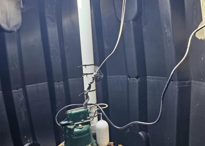 A green sump pump is installed on a concrete block inside a dark, cylindrical pit, with pipes and electrical wires connected. The pit appears to be partially filled with dirt and debris.