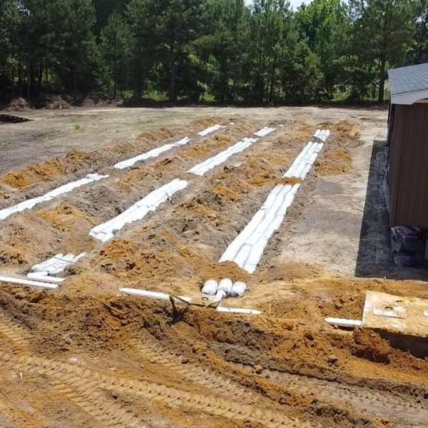 A construction site showing prepared ground with trenches and white support beams for a new building. Trees and a partially constructed structure are visible in the background.