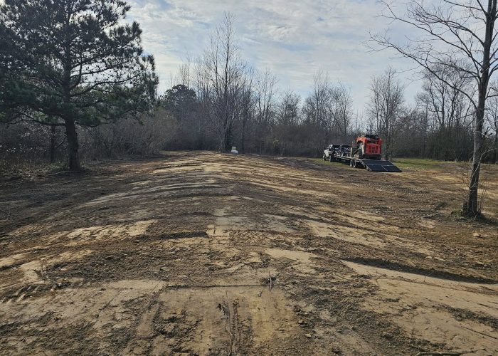 Cleared land with tire tracks leading into a wooded area, featuring a few bare trees and a construction vehicle in the background. The sky is partly cloudy.