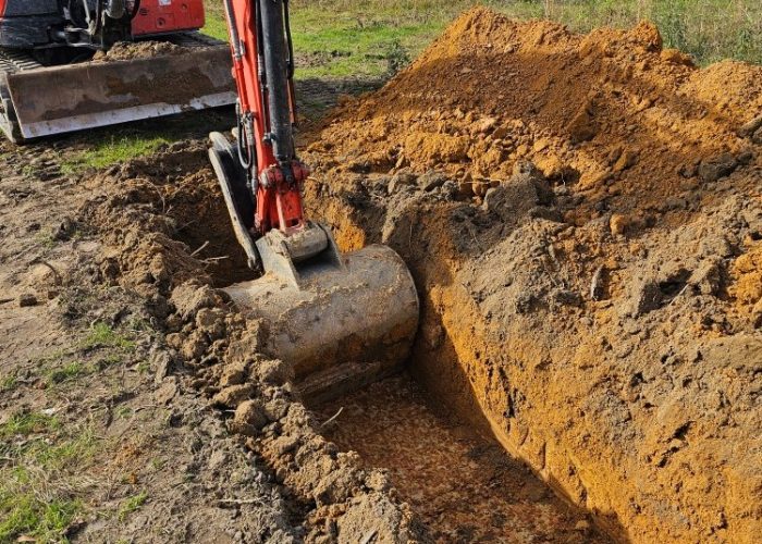 A backhoe excavator is digging a trench in a grassy area, with piles of orange soil beside it. The scene shows a clear sky and trees in the background.